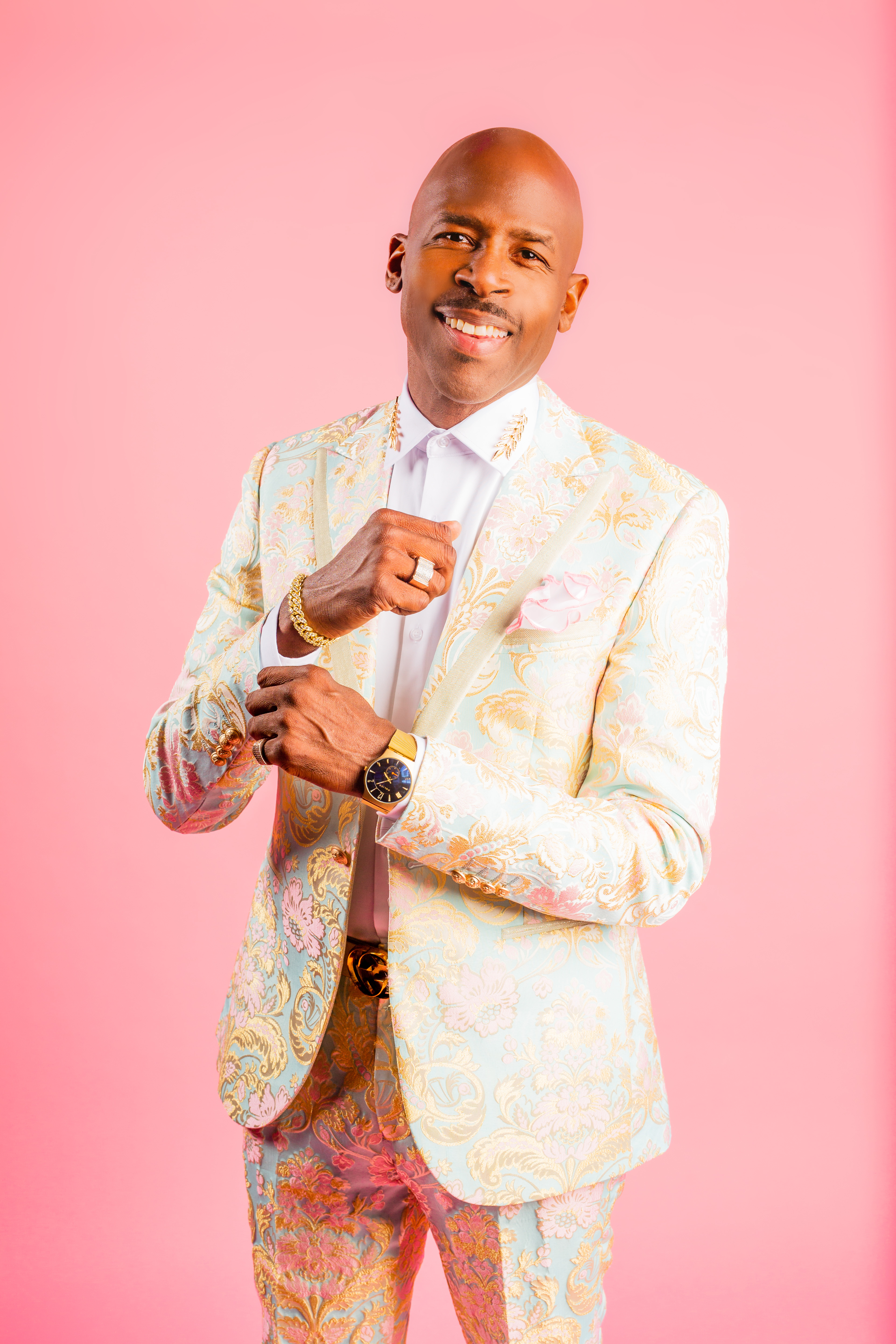 A smiling man in a colorful, patterned suit and gold jewelry stands against a pink background.