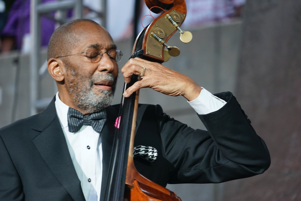 An elderly man with a long gray beard playing a cello on stage, wearing a tuxedo with a bow tie.