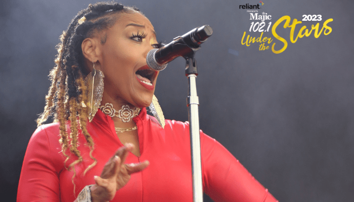 A woman with braided hair wearing a red outfit singing into a microphone on stage, with "Reliant Magic 102.1 Under the Stars 2023" text visible.