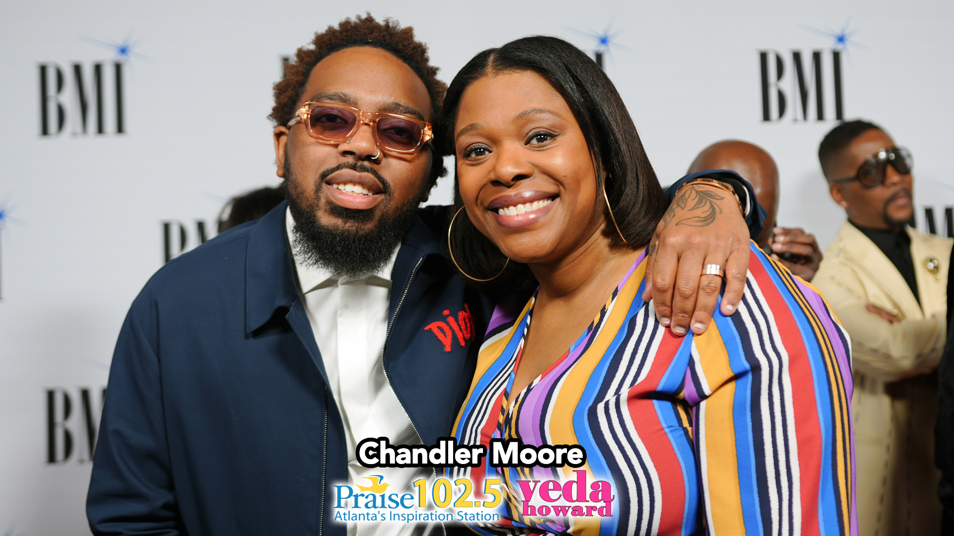 Two smiling people posing together at a BMI event, with the text "Chandler Moore" and "Praise 102.5 WYDA Atlanta's Inspiration Station" visible.