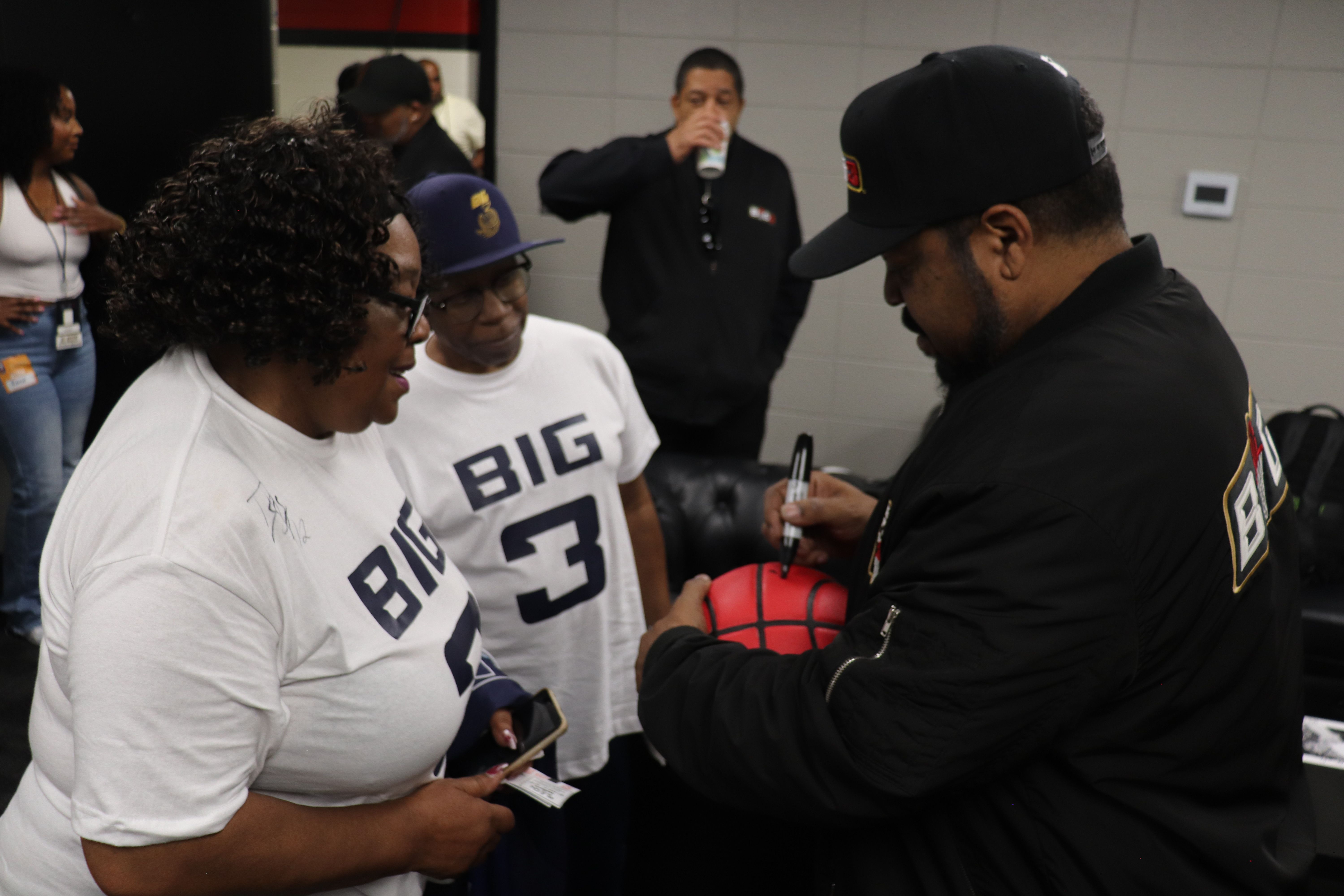 Several people in a room, one wearing a "Big 3" jersey, another wearing a black uniform, and others observing.