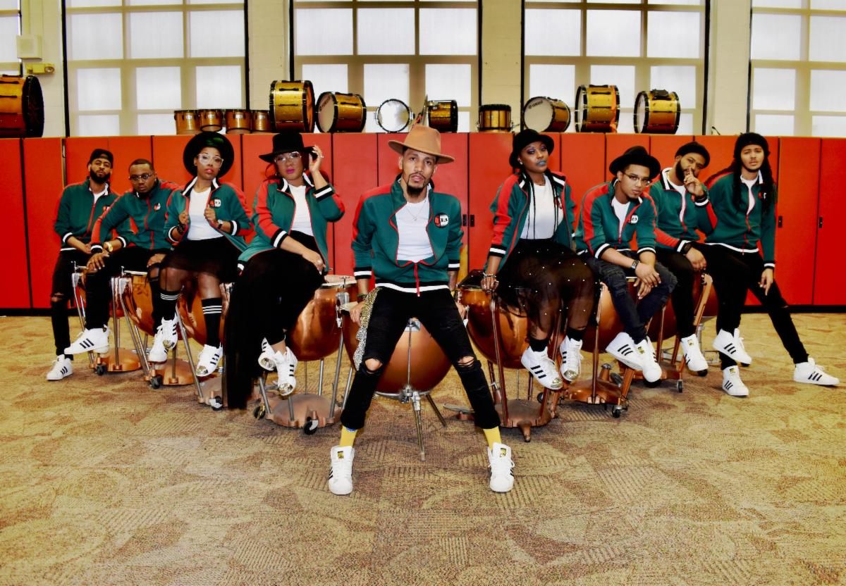 A group of people wearing green and black athletic uniforms, holding drums and other percussion instruments, posing in a room with large windows.