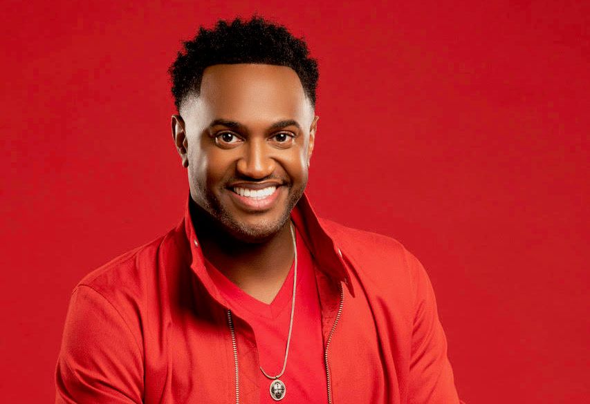 A smiling Black man wearing a red jacket and necklace against a bright red background.
