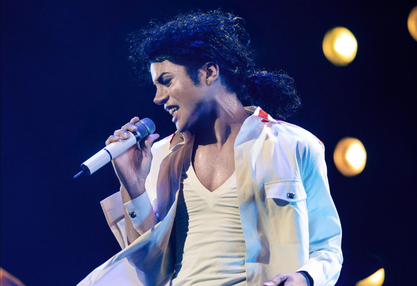 A man with curly hair performing on stage, holding a microphone and wearing a white jacket.