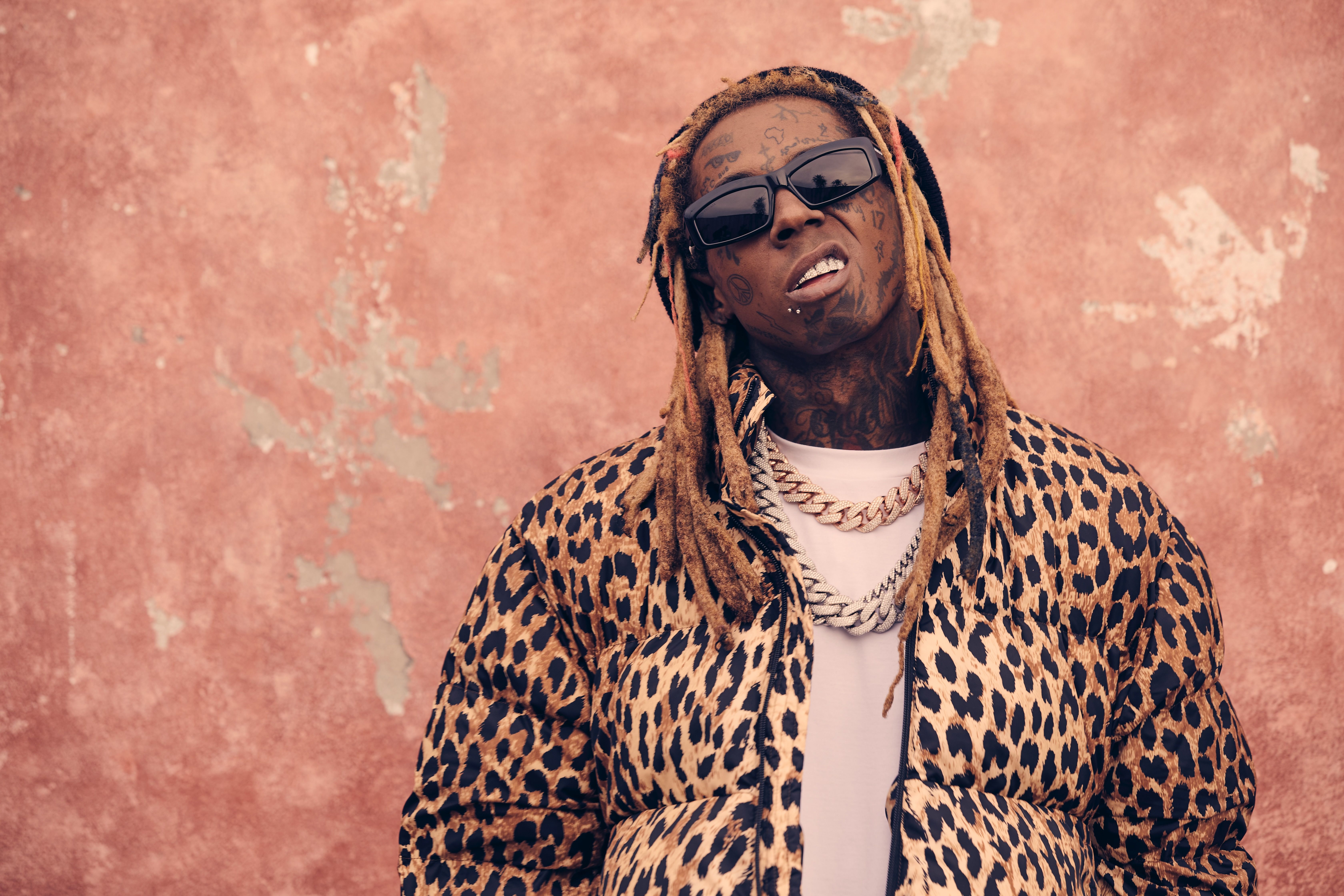 A man with dreadlocks wearing a leopard print jacket and sunglasses, smiling against a pink wall.