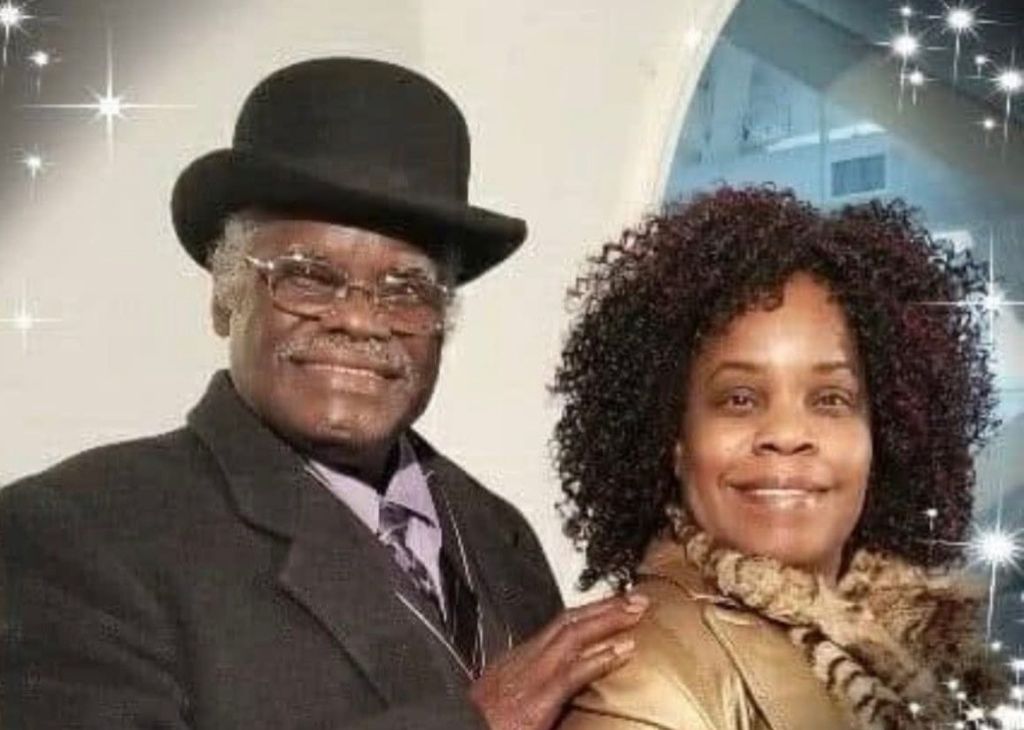 Two smiling African American adults, a man wearing a black bowler hat and a woman with curly hair, standing together in a formal setting.