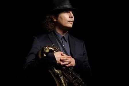 A person wearing a black hat and suit, holding a saxophone, standing in a dark setting.