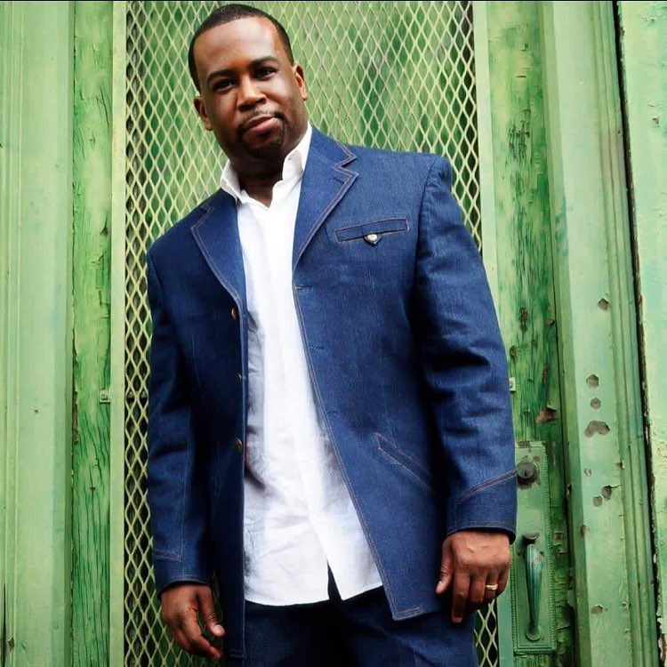 A well-dressed African American man in a blue suit standing in front of a green wooden lattice fence.