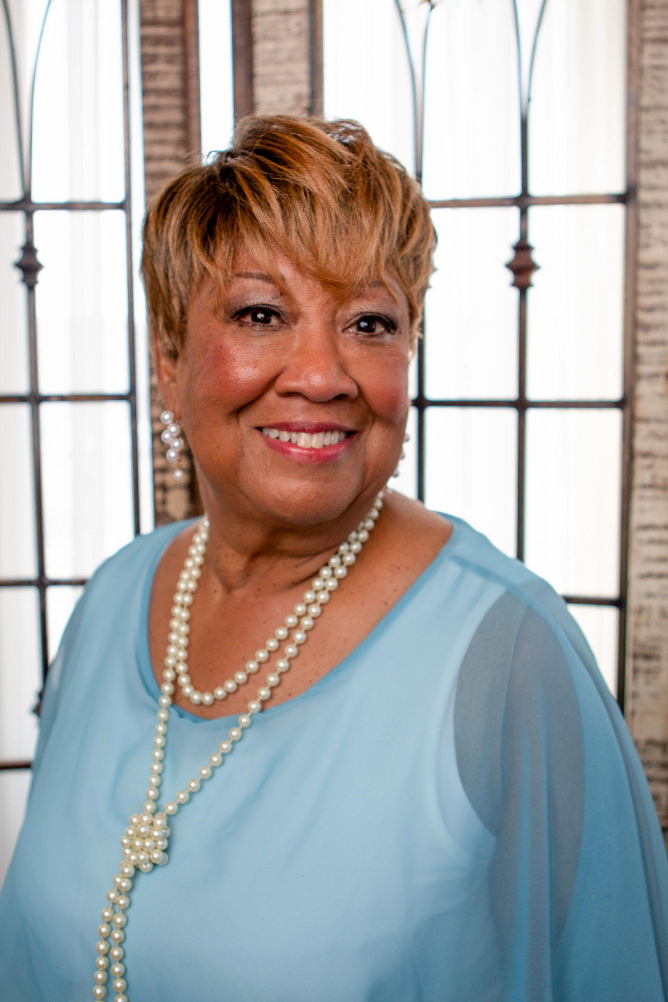 Smiling woman with short brown hair wearing a blue blouse and pearl necklace standing in front of a window.